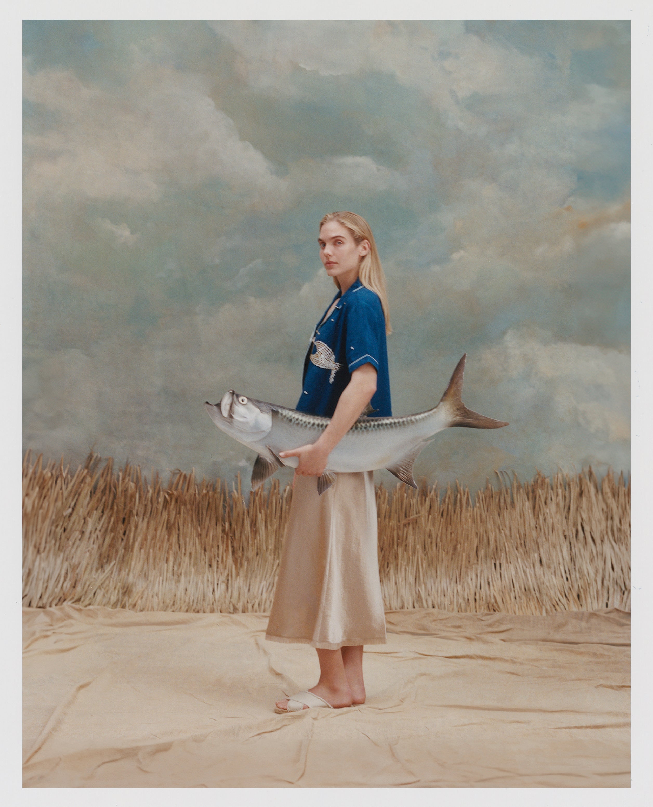 Female model with long blonde hair wearing the 'Teamwork' Cabana ~ Crop Cut while holding a model fish, and wearing a gold colored silk dress and white sandals, all shot on a background of texutred light blue and white with waist high beige grass.