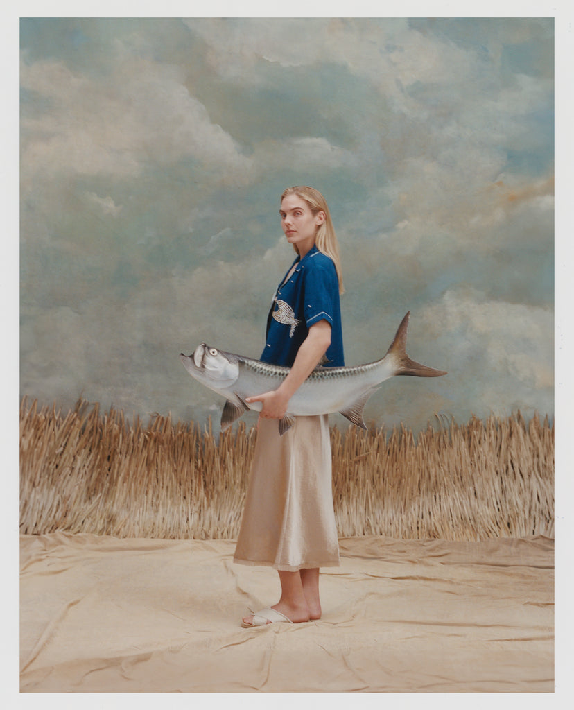 Female model with long blonde hair wearing the 'Teamwork' Cabana ~ Crop Cut while holding a model fish, and wearing a gold colored silk dress and white sandals, all shot on a background of texutred light blue and white with waist high beige grass.