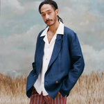 Male model with dreadlocks wearing the easy-going jacket in navy linen with a white shirt underneath with collars over the blazer, and red and black striped pants, shot on a light blue textured background with beige colored grass.