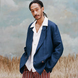 Male model with dreadlocks wearing the easy-going jacket in navy linen with a white shirt underneath with collars over the blazer, and red and black striped pants, shot on a light blue textured background with beige colored grass.