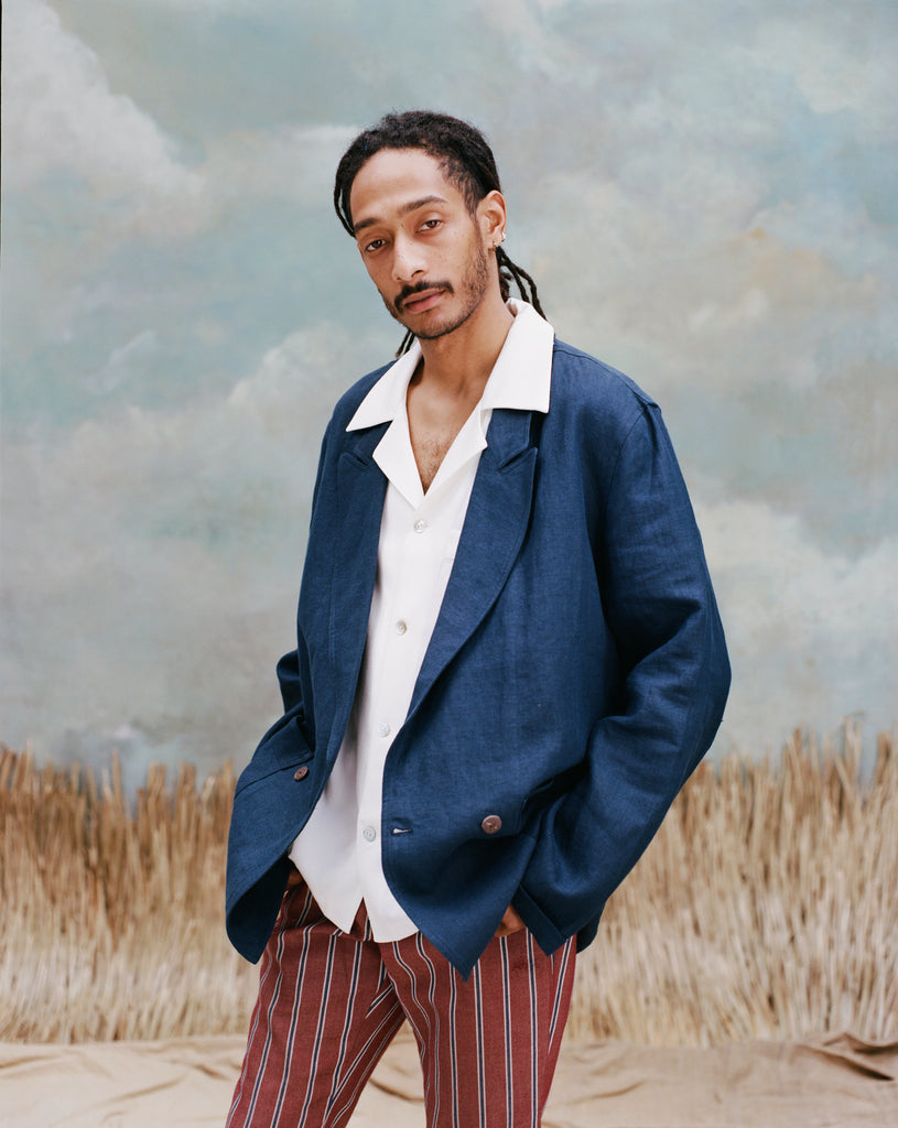 Male model with dreadlocks wearing the easy-going jacket in navy linen with a white shirt underneath with collars over the blazer, and red and black striped pants, shot on a light blue textured background with beige colored grass.