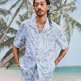 Male model with dreadlocks wearing 'Palm Fronds (Blue)' with the bottom button undone, alongside matching blue Palm Fronds shorts, shot on a background with palm trees and a sunny beach. 