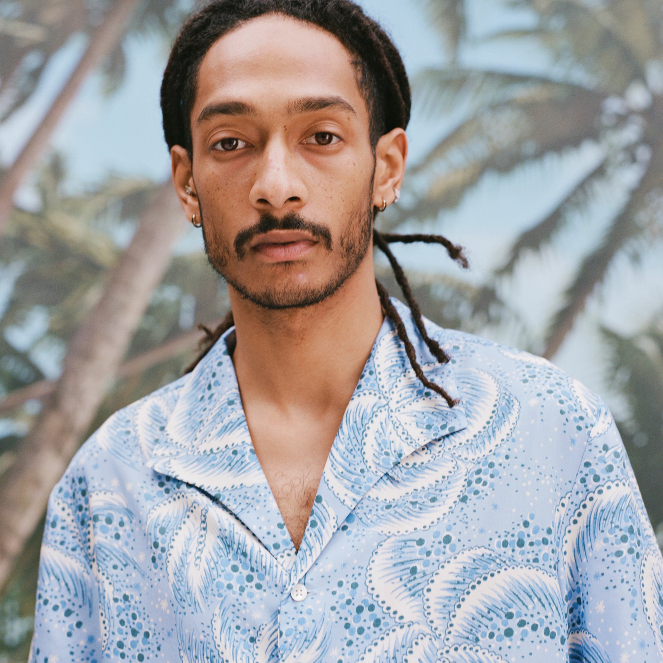 Close-up image of a male model with dreadlocks wearing 'Palm Fronds (Blue)' standing against a sunny background of palm trees.