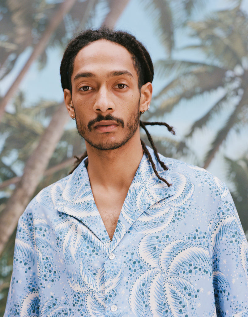 Close-up image of a male model with dreadlocks wearing 'Palm Fronds (Blue)' standing against a sunny background of palm trees.