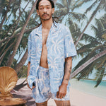 Male model with dreadlocks wearing an unbuttoned 'Palm Fronds (Blue)' with matching Palm Fronds (blue) shorts, shot on a background of palm trees and a sunny beach.