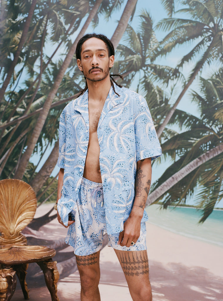 Male model with dreadlocks wearing an unbuttoned 'Palm Fronds (Blue)' with matching Palm Fronds (blue) shorts, shot on a background of palm trees and a sunny beach.