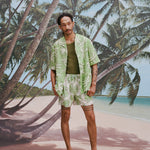 Male model with dreadlocks wearing an unbuttoned 'Palm Fronds (green)' with a dark green tank underneath and matching 'Palm Fronds' (Green), all shot on a background with palm trees and a sunny beach.