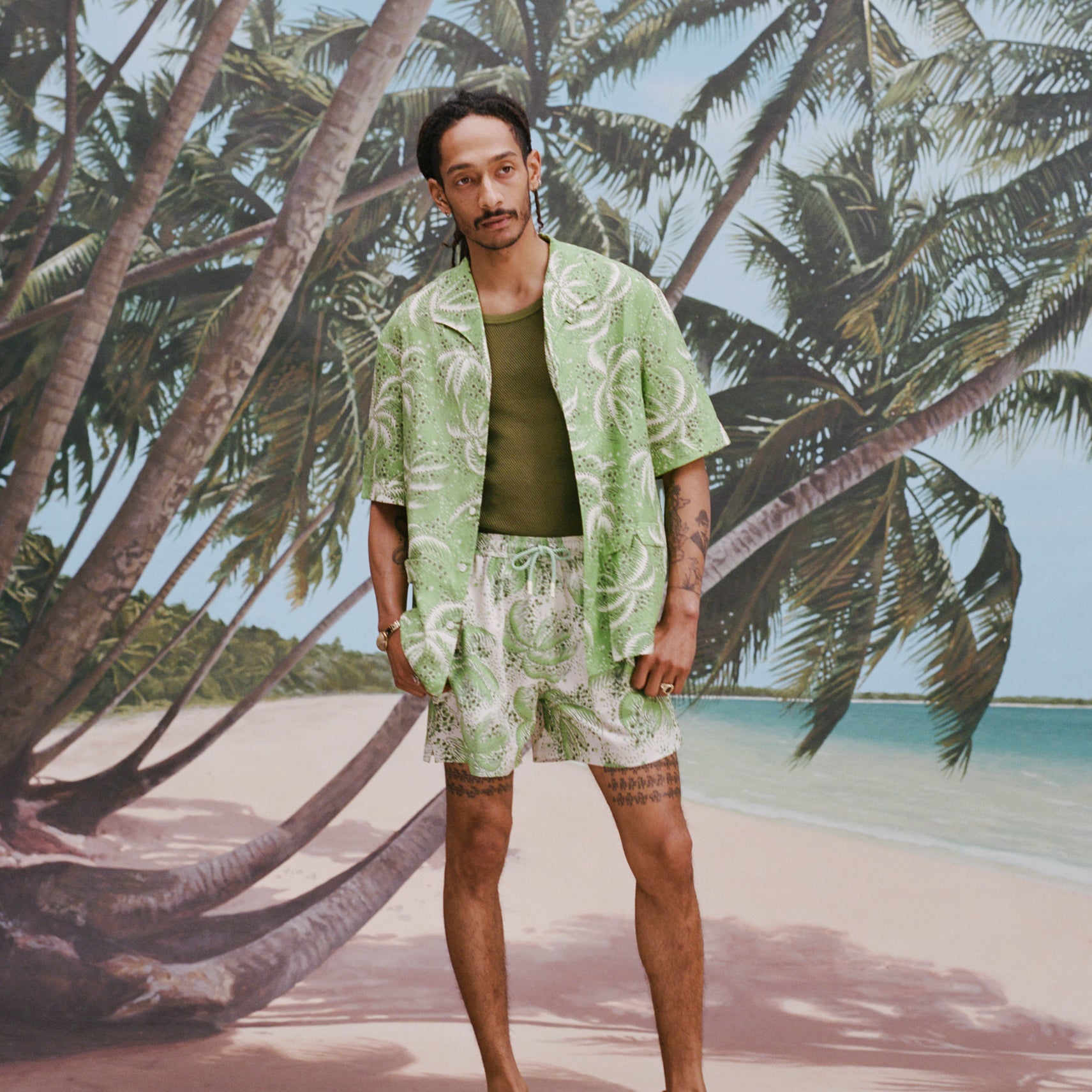 Male model with dreadlocks wearing an unbuttoned 'Palm Fronds (green)' with a dark green tank underneath and matching 'Palm Fronds' (Green), all shot on a background with palm trees and a sunny beach.