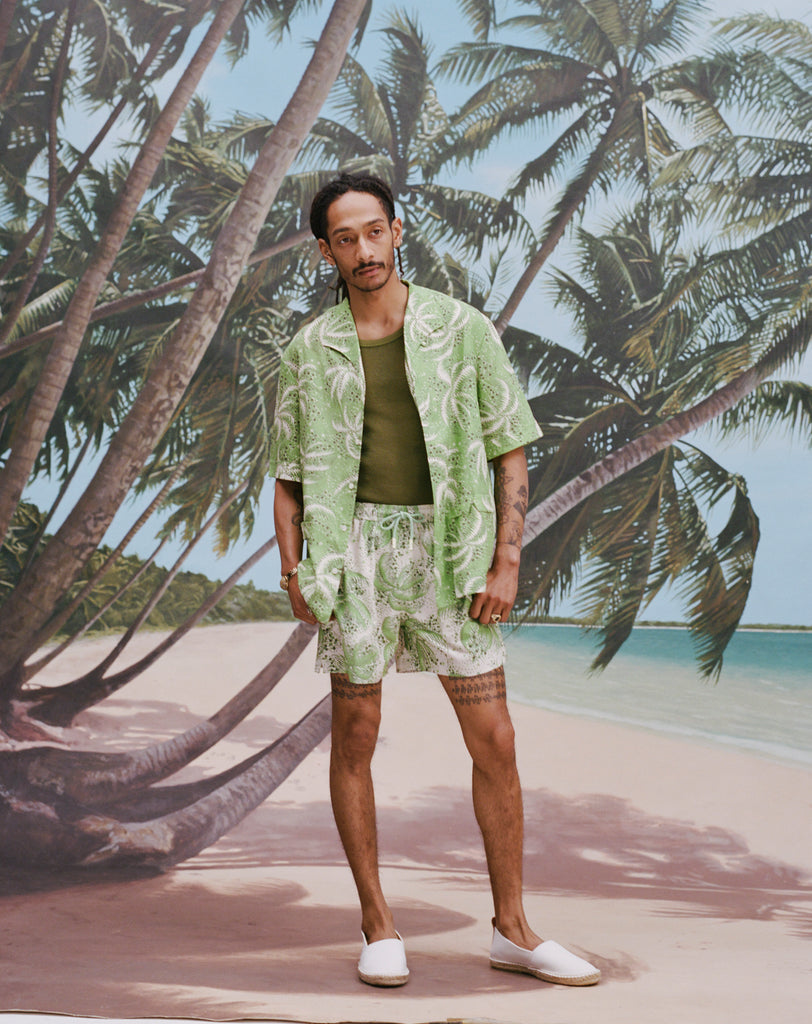 Male model with dreadlocks wearing an unbuttoned 'Palm Fronds (green)' with a dark green tank underneath and matching 'Palm Fronds' (Green), all shot on a background with palm trees and a sunny beach.