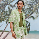 Male model with dreadlocks wearing an unbuttoned 'Palm Fronds (Green)' with a dark green tank and matching 'Palm Fronds (green)' shorts and a gold watch on his wrist, all shot on background with palm trees and a sunny beach.