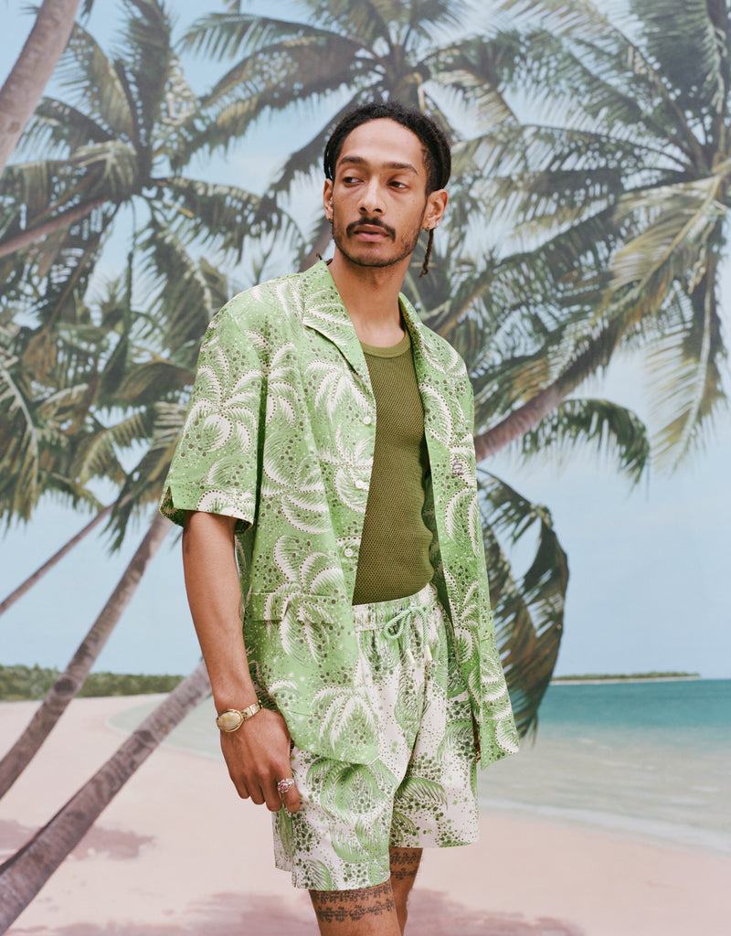 Male model with dreadlocks wearing an unbuttoned 'Palm Fronds (Green)' with a dark green tank and matching 'Palm Fronds (green)' shorts and a gold watch on his wrist, all shot on background with palm trees and a sunny beach.