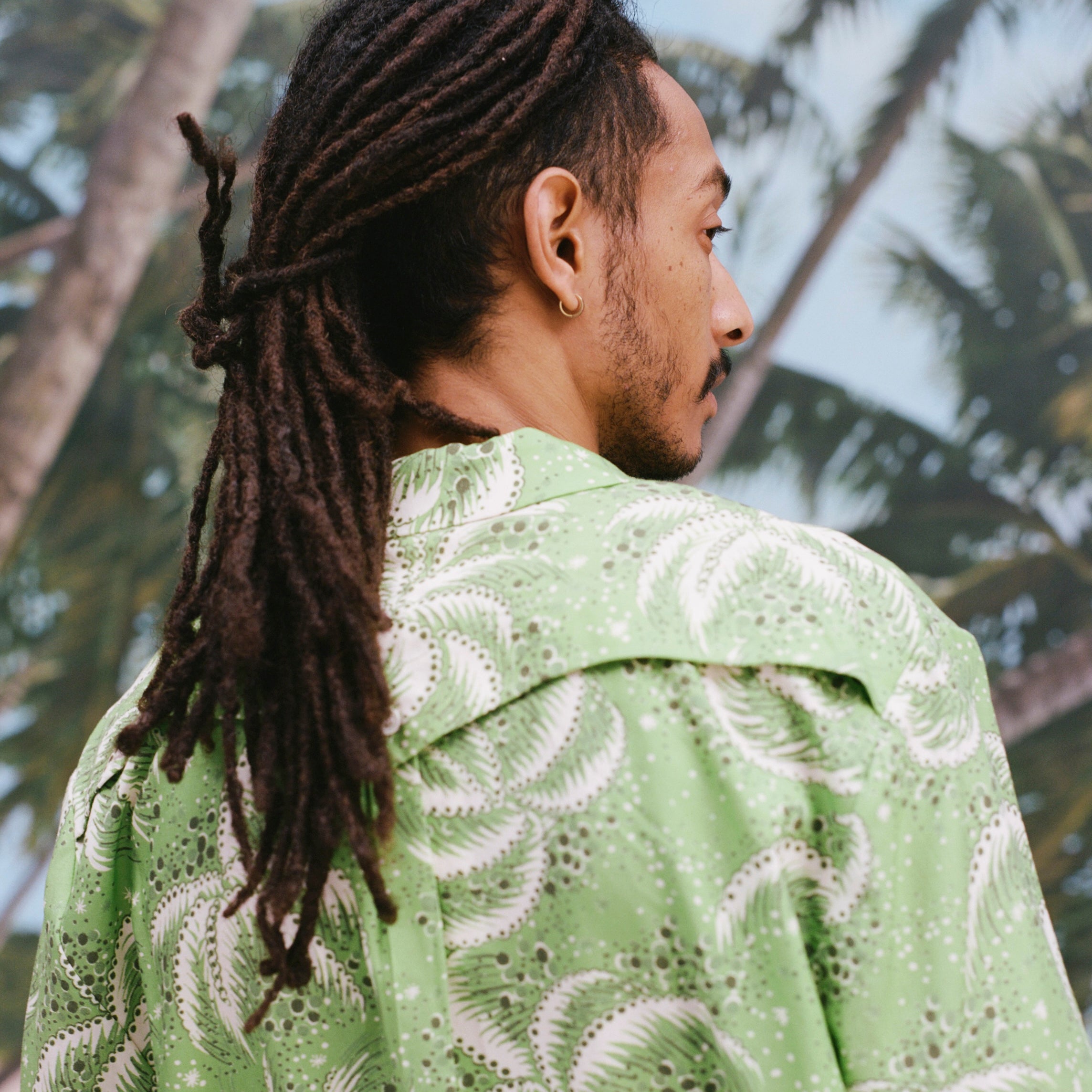 Backwards-facing image of a male model with dreadlocks wearing 'Palm Fronds (green),' shot on a background with blue skies and palm trees.