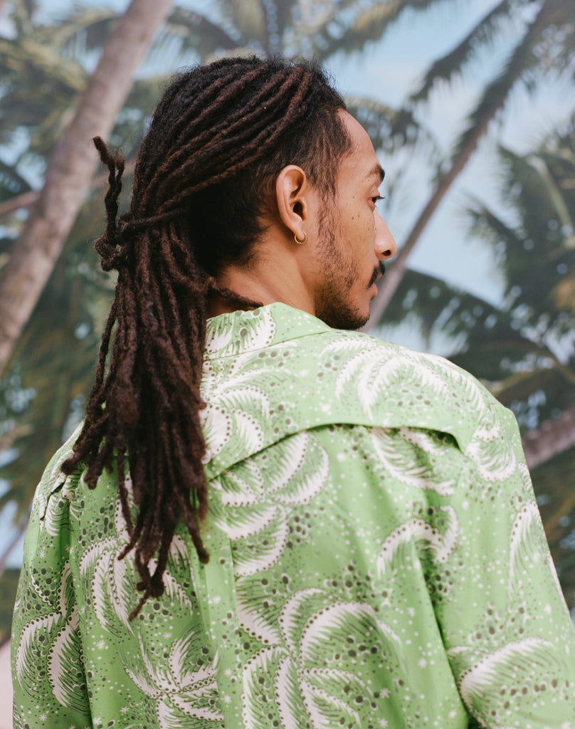Backwards-facing image of a male model with dreadlocks wearing 'Palm Fronds (green),' shot on a background with blue skies and palm trees.