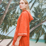 Female model with long blonde hair turned to the side with her face angled toward the camera wearing a 'Scallop' Beach Towel Top showcasing the fringed hems on sleeves and bottom, while wearing a mesh skirt against a background of palm trees, blue skies, and a beach.