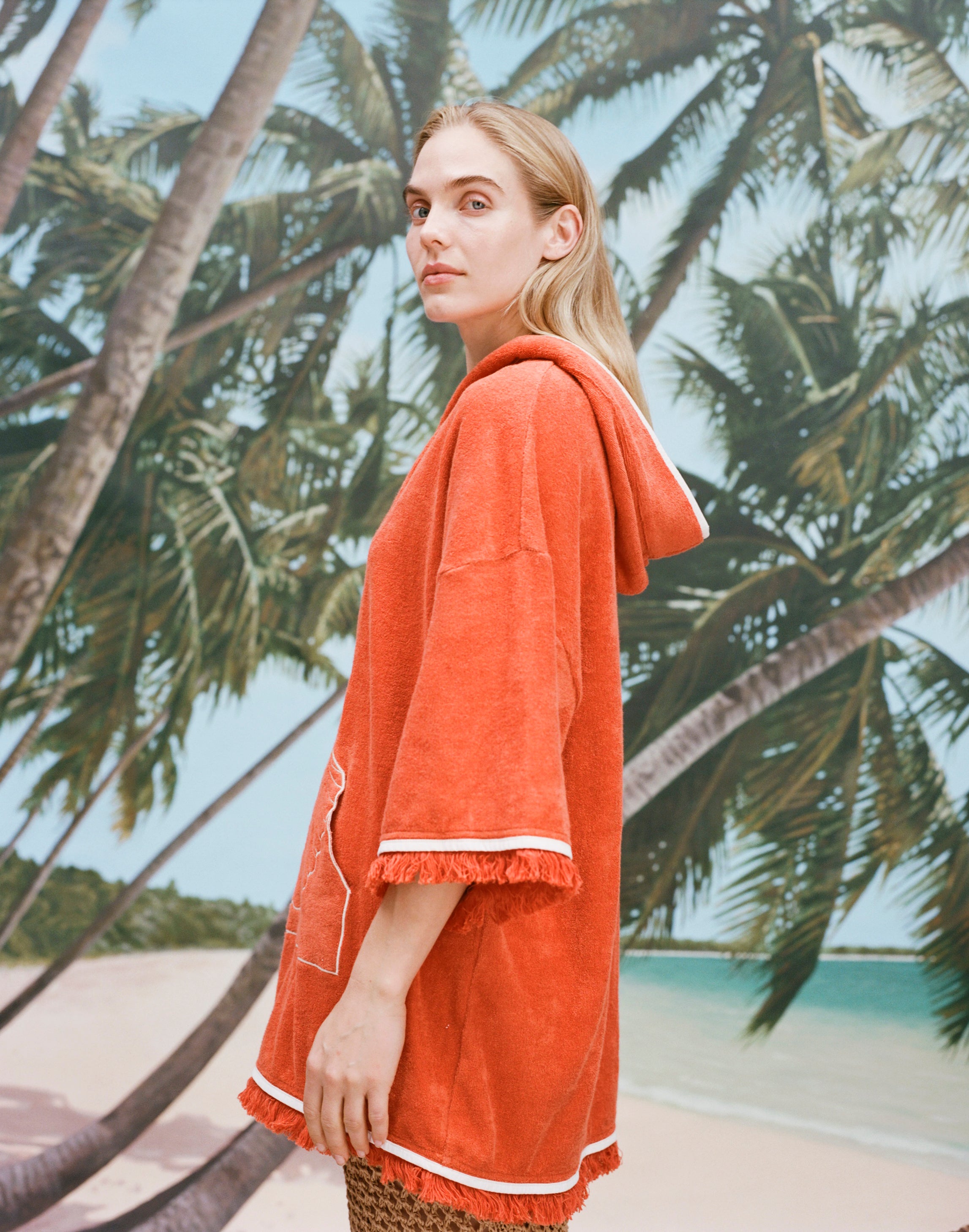 Female model with long blonde hair turned to the side with her face angled toward the camera wearing a 'Scallop' Beach Towel Top showcasing the fringed hems on sleeves and bottom, while wearing a mesh skirt against a background of palm trees, blue skies, and a beach.