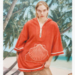 Female model with long blonde hair wearing the 'Scallop' Beach Towel top in Coral coloring against a background of palm trees, blue skies, and a beach, with a mesh skirt.