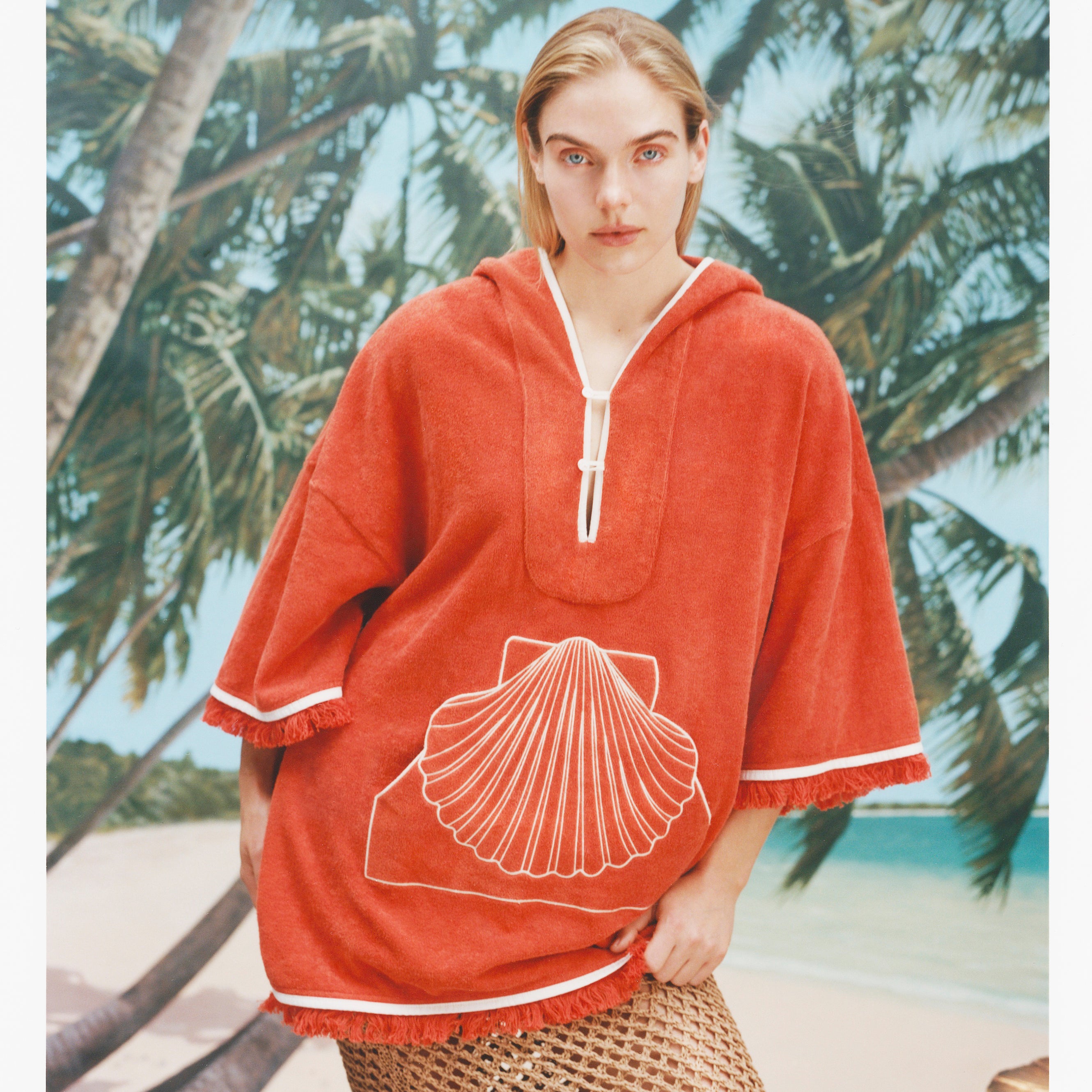 Female model with long blonde hair wearing the 'Scallop' Beach Towel top in Coral coloring against a background of palm trees, blue skies, and a beach, with a mesh skirt.
