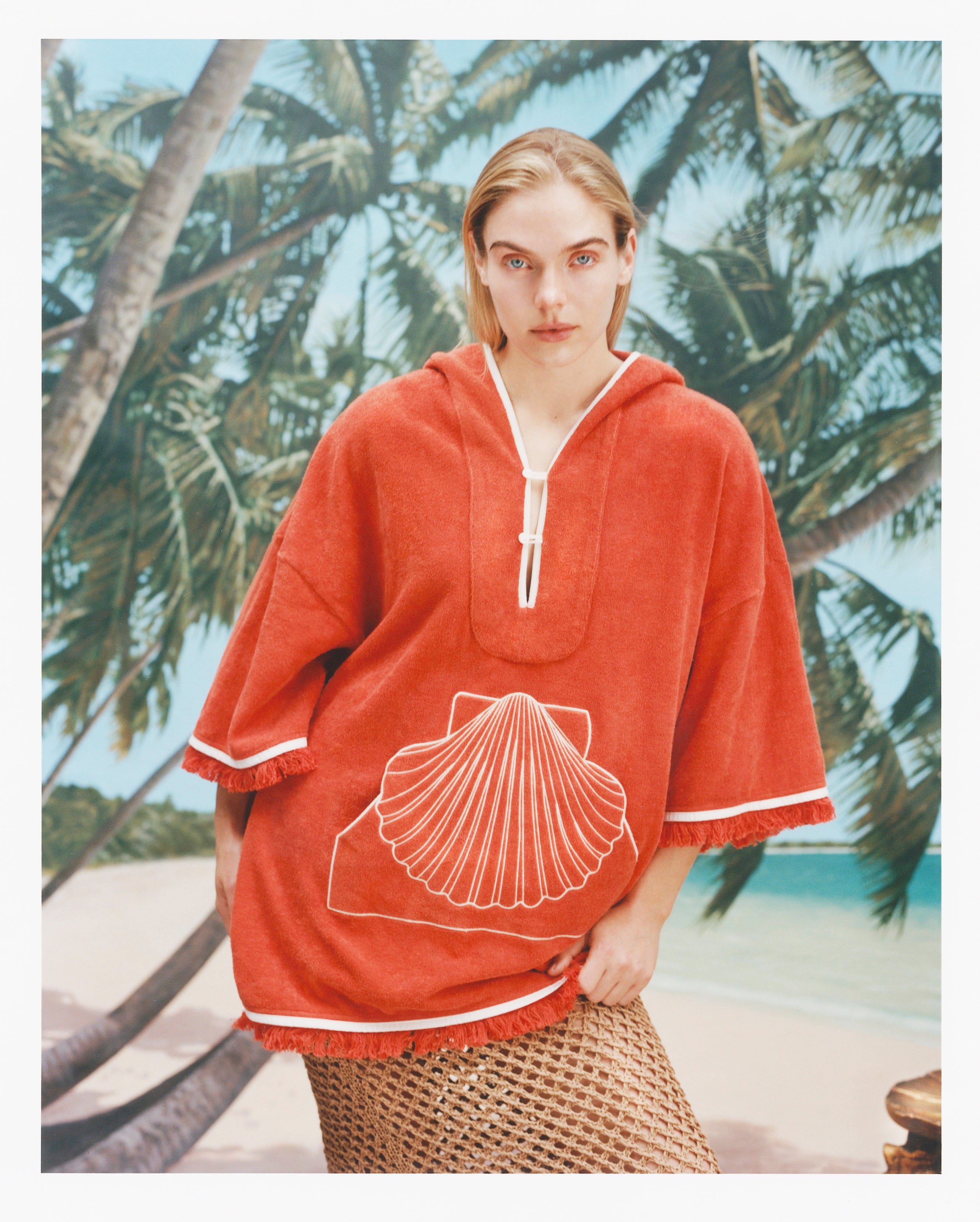 Female model with long blonde hair wearing the 'Scallop' Beach Towel top in Coral coloring against a background of palm trees, blue skies, and a beach, with a mesh skirt.