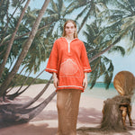 Female model with long blonde hair wearing the 'Scallop' Beach Towel Top and a mesh full-length skirt in khaki coloring with white sandals, shot on a background of a sunny beach with palm trees and a bamboo chair.