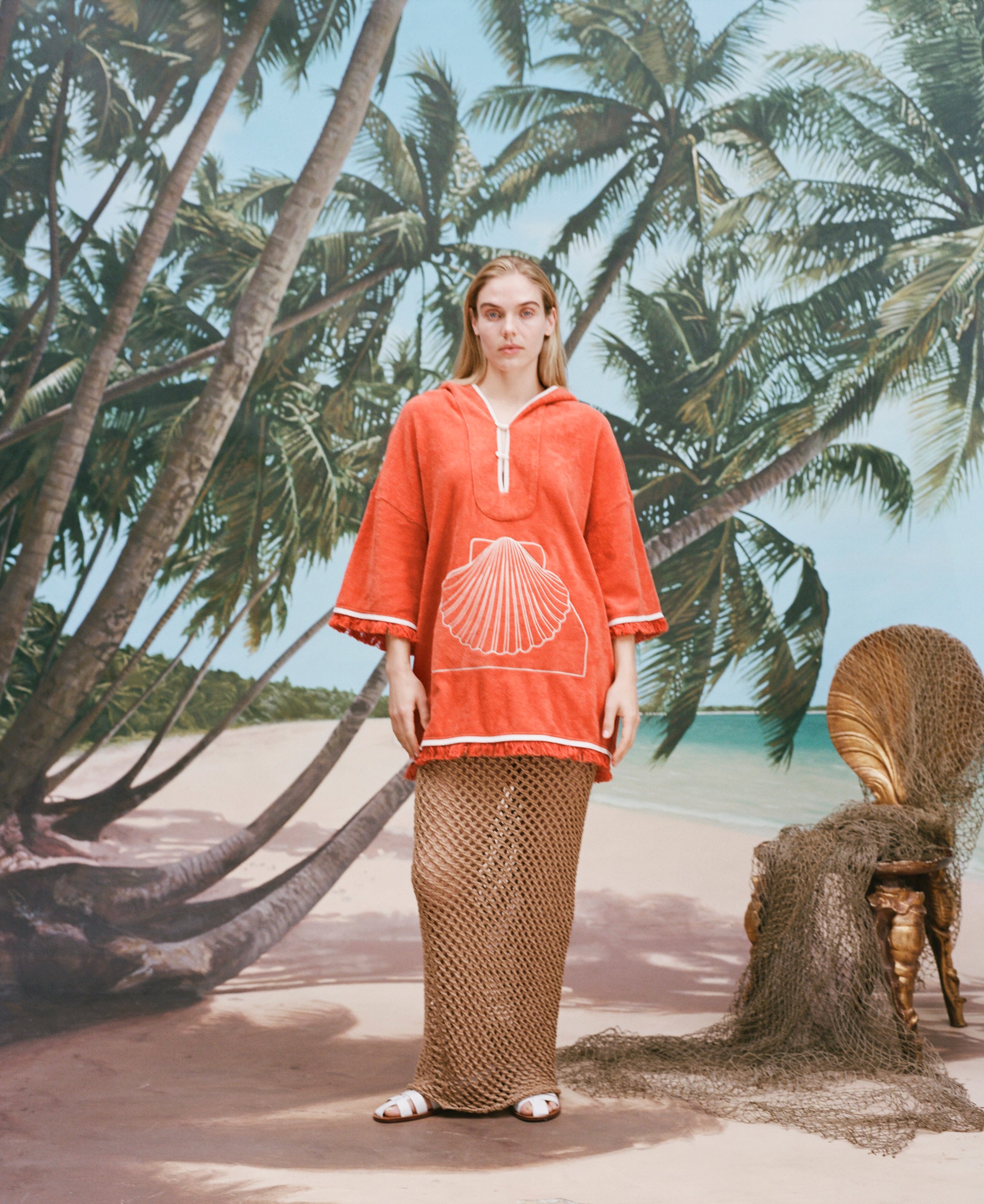 Female model with long blonde hair wearing the 'Scallop' Beach Towel Top and a mesh full-length skirt in khaki coloring with white sandals, shot on a background of a sunny beach with palm trees and a bamboo chair.