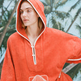 Female model with long blonde hair wearing the 'Scallop' Beach Towel Top with the hood over her head, and one hand on her waist, shot on a background of blue skies and palm trees.