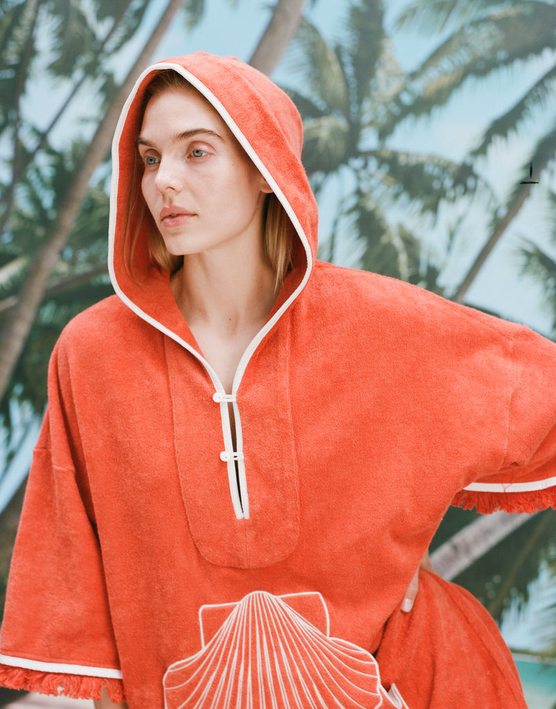 Female model with long blonde hair wearing the 'Scallop' Beach Towel Top with the hood over her head, and one hand on her waist, shot on a background of blue skies and palm trees.