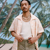 Male model with dreadlocks wearing the Voyager Shirt in coral seersucker unbuttoned with a white tank top and light green pants, shot on a background with palm trees, blue skies, and a beach.