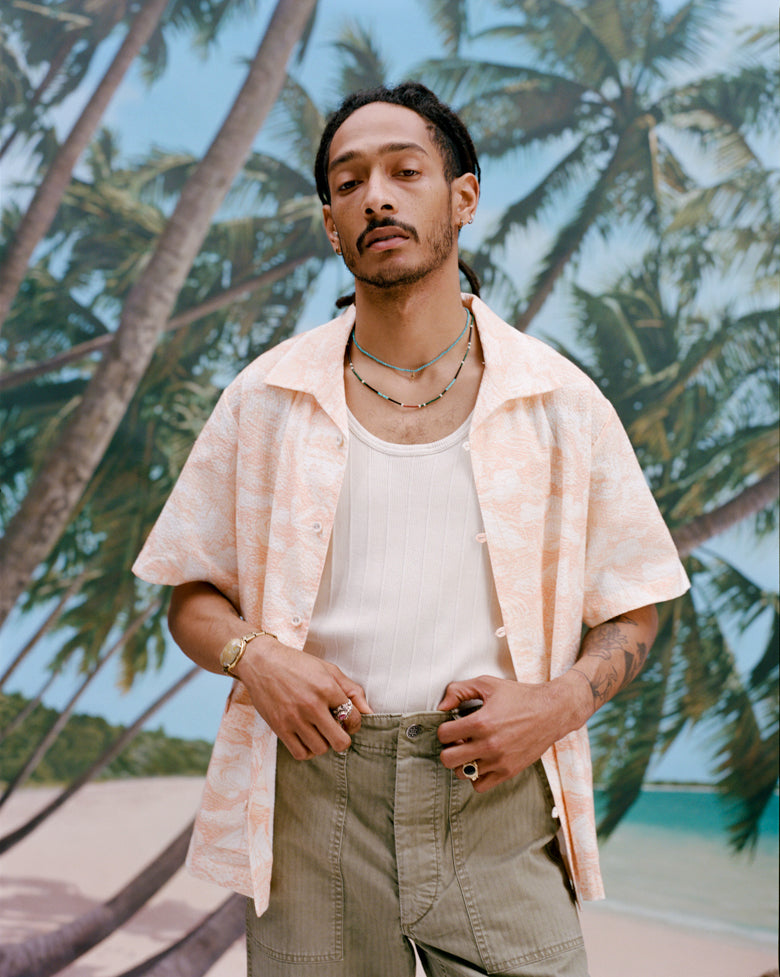 Male model with dreadlocks wearing the Voyager Shirt in coral seersucker unbuttoned with a white tank top and light green pants, shot on a background with palm trees, blue skies, and a beach.