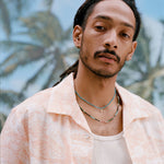 Male model with dreadlocks wearing the Voyager Shirt in Coral Seersucker unbuttoned with a white tank top underneath, shot on a background with palm fronds and blue skies.
