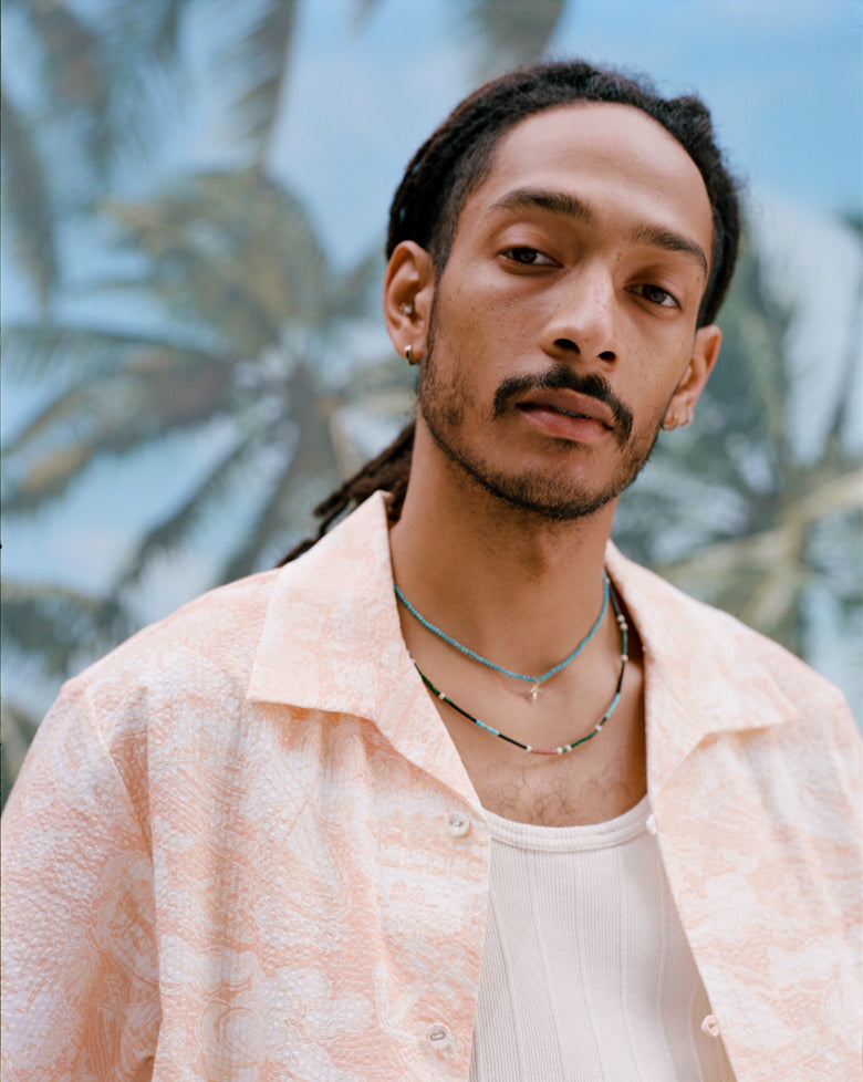 Male model with dreadlocks wearing the Voyager Shirt in Coral Seersucker unbuttoned with a white tank top underneath, shot on a background with palm fronds and blue skies.