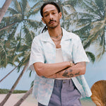 Male model with dreadlocks has his arms crossed while wearing the 'Voyager Shirt' in acqua seersucker unbuttoned with a white tank top underneath with light blue shorts, shot on a background of palm trees, and a beach. 