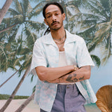 Male model with dreadlocks has his arms crossed while wearing the 'Voyager Shirt' in acqua seersucker unbuttoned with a white tank top underneath with light blue shorts, shot on a background of palm trees, and a beach. 