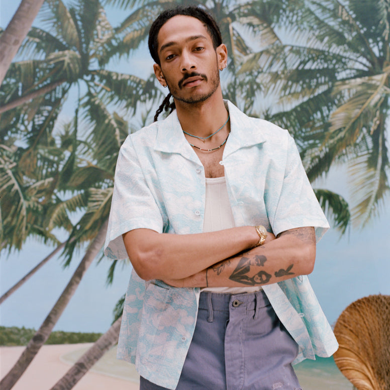 Male model with dreadlocks has his arms crossed while wearing the 'Voyager Shirt' in acqua seersucker unbuttoned with a white tank top underneath with light blue shorts, shot on a background of palm trees, and a beach. 