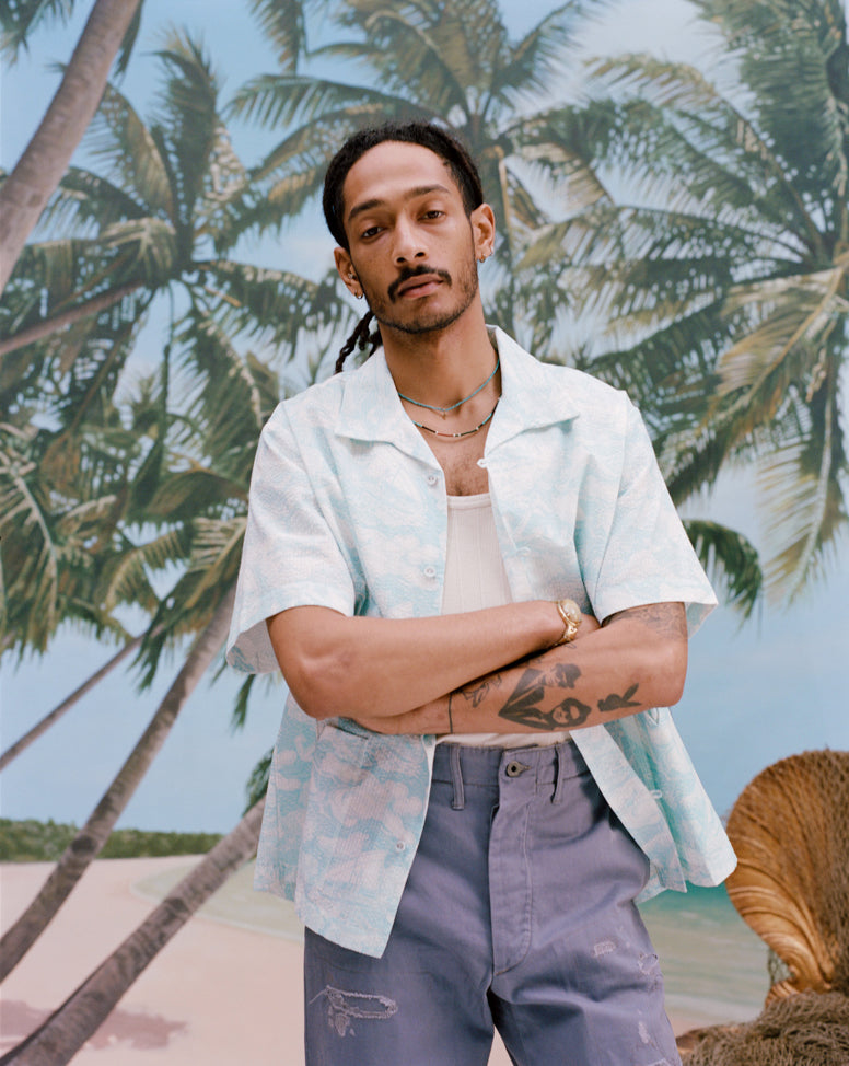 Male model with dreadlocks has his arms crossed while wearing the 'Voyager Shirt' in acqua seersucker unbuttoned with a white tank top underneath with light blue shorts, shot on a background of palm trees, and a beach. 