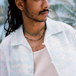 Male model with dreadlocks wearing the 'Voyager Shirt' in aqua seersucker unbuttoned with a white tank top beneath and two coral colored necklaces on, shot on a background of blue skies and palm fronds.