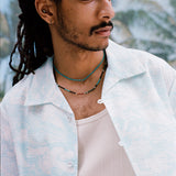 Male model with dreadlocks wearing the 'Voyager Shirt' in aqua seersucker unbuttoned with a white tank top beneath and two coral colored necklaces on, shot on a background of blue skies and palm fronds.