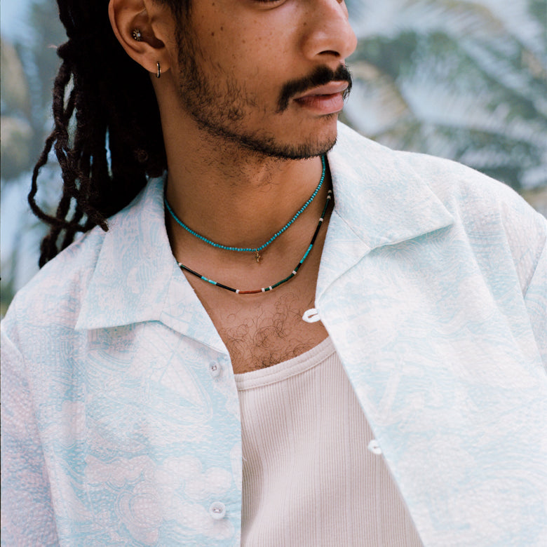 Male model with dreadlocks wearing the 'Voyager Shirt' in aqua seersucker unbuttoned with a white tank top beneath and two coral colored necklaces on, shot on a background of blue skies and palm fronds.