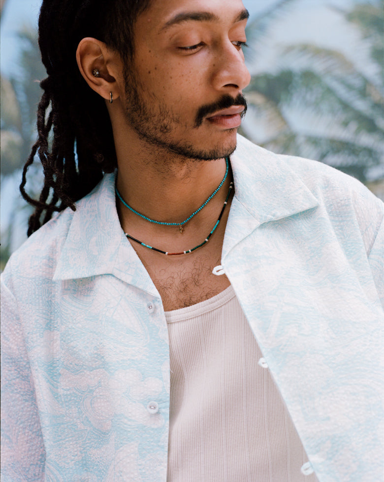 Male model with dreadlocks wearing the 'Voyager Shirt' in aqua seersucker unbuttoned with a white tank top beneath and two coral colored necklaces on, shot on a background of blue skies and palm fronds.