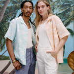 Male model with dreadlocks wearing the 'Voyager Shirt' in aqua seersucker unbuttoned with a white tank top underneath and indigo jeans with a female model with long blonde hair standing beside him wearing the 'Voyager Shirt' in Coral seersucker with a white shirt underneath and white pants, shot on a background with a beach, blue skies, and palm trees.