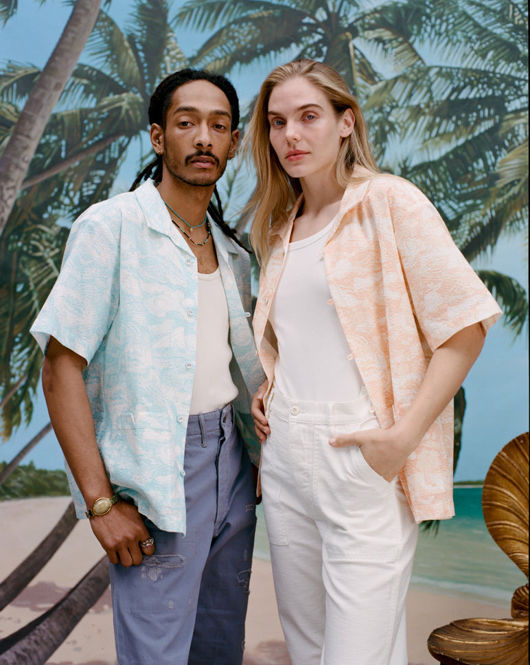 Male model with dreadlocks wearing the 'Voyager Shirt' in aqua seersucker unbuttoned with a white tank top underneath and indigo jeans with a female model with long blonde hair standing beside him wearing the 'Voyager Shirt' in Coral seersucker with a white shirt underneath and white pants, shot on a background with a beach, blue skies, and palm trees.