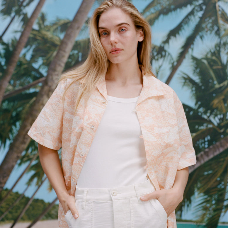 Female model with long-blonde hair wearing the 'Voyager Shirt' in coral seersucker unbuttoned with a white shirt underneath and white pants,  shot on a background with palm trees, blue skies, and a beach.