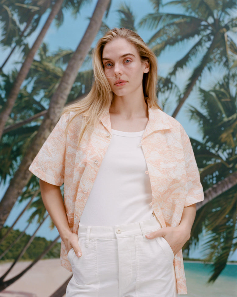 Female model with long-blonde hair wearing the 'Voyager Shirt' in coral seersucker unbuttoned with a white shirt underneath and white pants,  shot on a background with palm trees, blue skies, and a beach.
