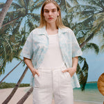Female model with long blonde hair wearing the Voyager Shirt in aqua seersucker unbuttoned with a white shirt underneath that is tucked into white pants, shot on a background with a beach, blue skies, and palm trees.