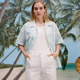 Female model with long blonde hair wearing the Voyager Shirt in aqua seersucker unbuttoned with a white shirt underneath that is tucked into white pants, shot on a background with a beach, blue skies, and palm trees.