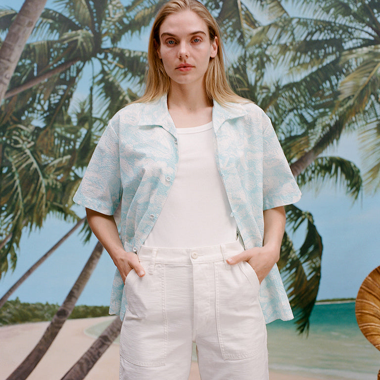 Female model with long blonde hair wearing the Voyager Shirt in aqua seersucker unbuttoned with a white shirt underneath that is tucked into white pants, shot on a background with a beach, blue skies, and palm trees.