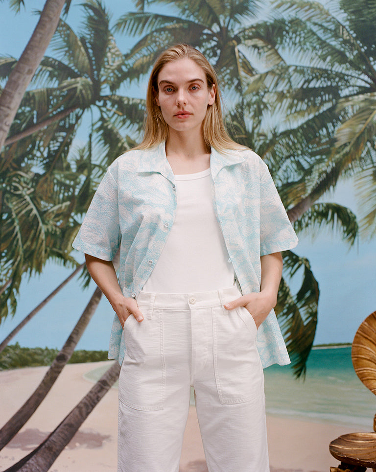 Female model with long blonde hair wearing the Voyager Shirt in aqua seersucker unbuttoned with a white shirt underneath that is tucked into white pants, shot on a background with a beach, blue skies, and palm trees.