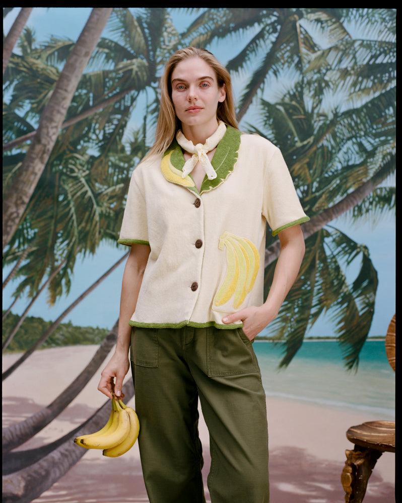 A woman wearing a cream-colored short-sleeve shirt with green trim and a large banana applique on the front, green pants, and a light scarf tied around her neck. She holds a bunch of bananas in one hand.