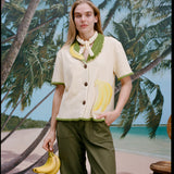 A woman wearing a cream-colored short-sleeve shirt with green trim and a large banana applique on the front, green pants, and a light scarf tied around her neck. She holds a bunch of bananas in one hand.