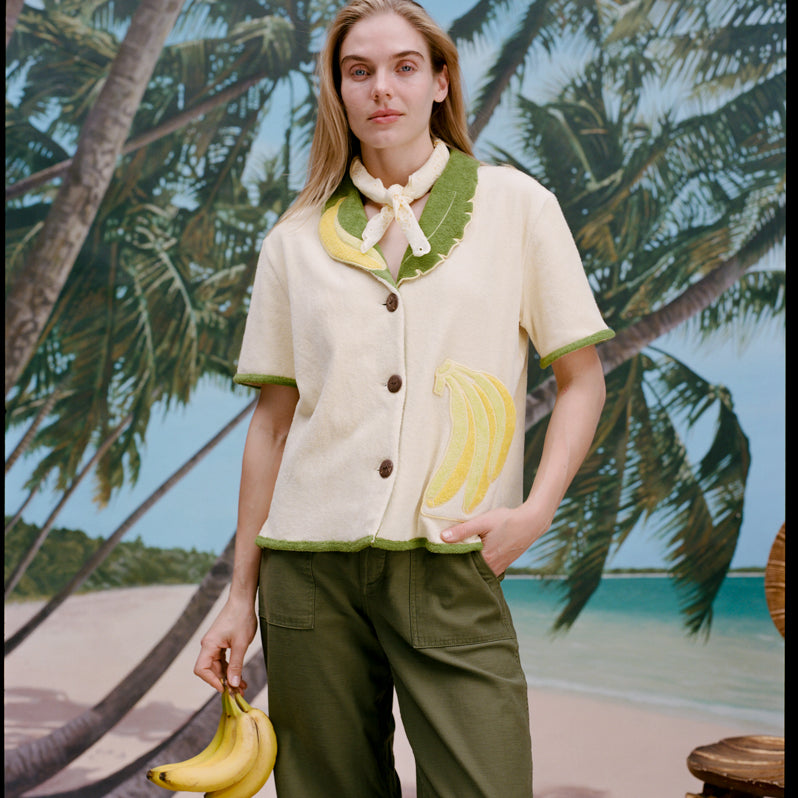 A woman wearing a cream-colored short-sleeve shirt with green trim and a large banana applique on the front, green pants, and a light scarf tied around her neck. She holds a bunch of bananas in one hand.