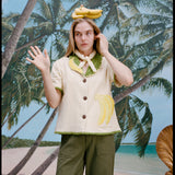 A woman wearing a cream-colored short-sleeve shirt with green trim and a large banana applique on the front, green pants, and a light scarf tied around her neck. She has a bunch of bananas sitting on top of her head. 